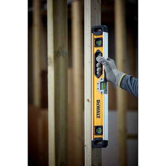A DEWALT spirit level being held vertically against a wooden post by a gloved hand, used to check alignment during construction.