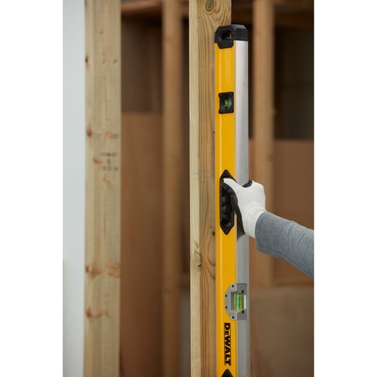 A person wearing a glove is holding a yellow DEWALT spirit level against a vertical wooden beam in a construction setting.