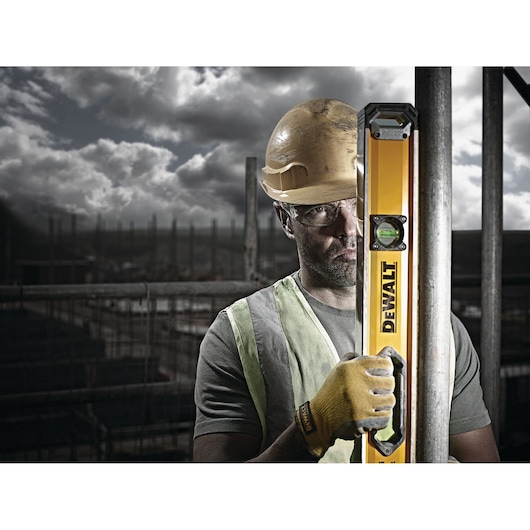 A construction worker wearing a safety helmet, reflective vest, and yellow gloves is holding a yellow DEWALT spirit level against a metal pole at a construction site. The background shows a cloudy sky and building scaffolding.