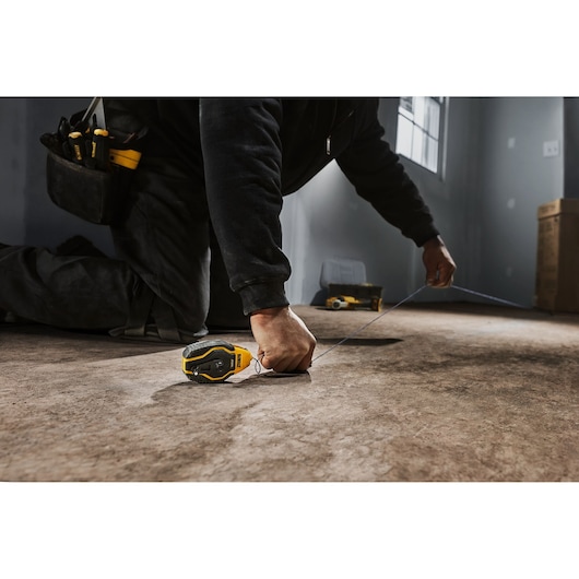 A person kneeling on the floor uses a DEWALT chalk line tool (DWHT47258L) to mark a straight line on a surface in a partially finished room. The individual has a tool belt and is shown pulling the chalk line tight.
