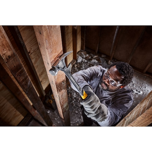 A worker wearing gloves uses a DEWALT claw hammer (SKU: DWHT51008) to drive a nail into a wooden beam inside a construction site. The worker's face is blurred for privacy.