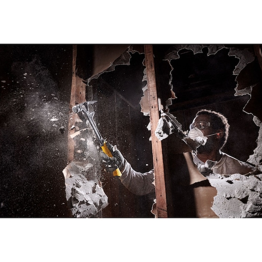 A person with a blurred face is using a DEWALT hammer to break through a drywall, creating dust and debris. The hammer has a yellow handle and is held with a protective glove.