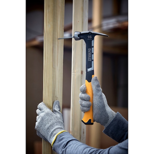 A person wearing grey gloves is using a DEWALT hammer with a yellow and black handle to drive a nail into a wooden beam.