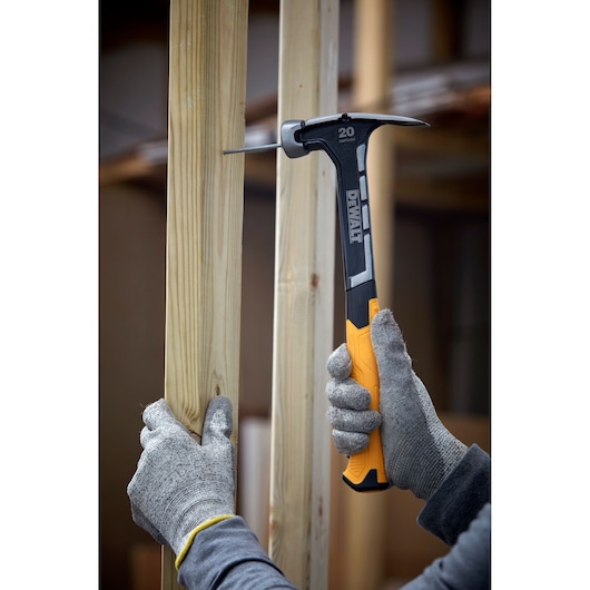 A pair of gloved hands using a DEWALT 20 oz hammer with a yellow handle to drive a nail into a vertical wooden beam.