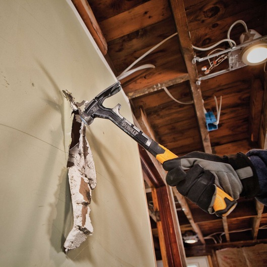 A person wearing work gloves uses a DEWALT hammer to break through a drywall in a construction or renovation setting, with exposed wooden ceiling beams and electrical wiring visible.