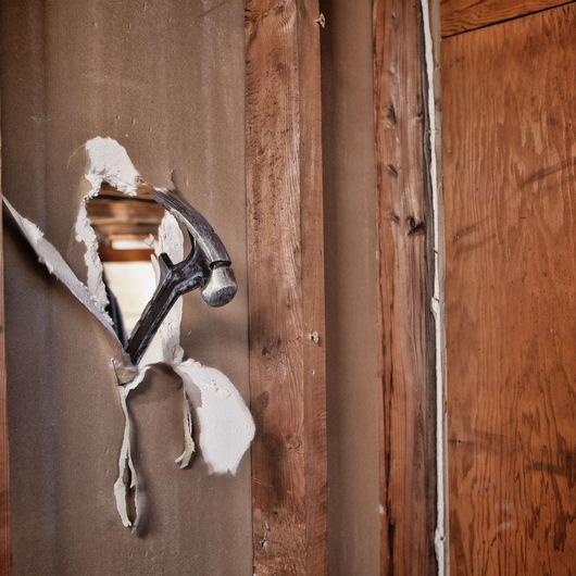 A hammer breaking through drywall, with its head protruding from a hole in the wall amidst exposed wooden framing.
