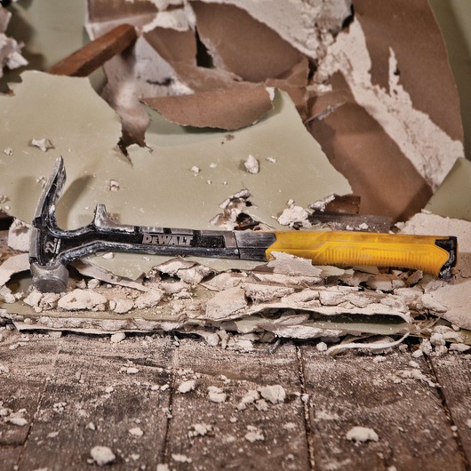 A DEWALT claw hammer with a yellow handle lies on broken drywall and debris on a construction site floor.
