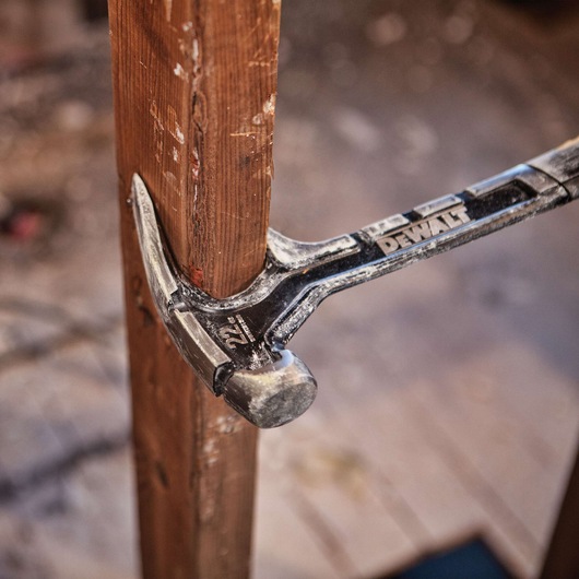 A DEWALT claw hammer with a 22-ounce marking is shown in use, pulling a nail from a wooden board in a construction or renovation setting.