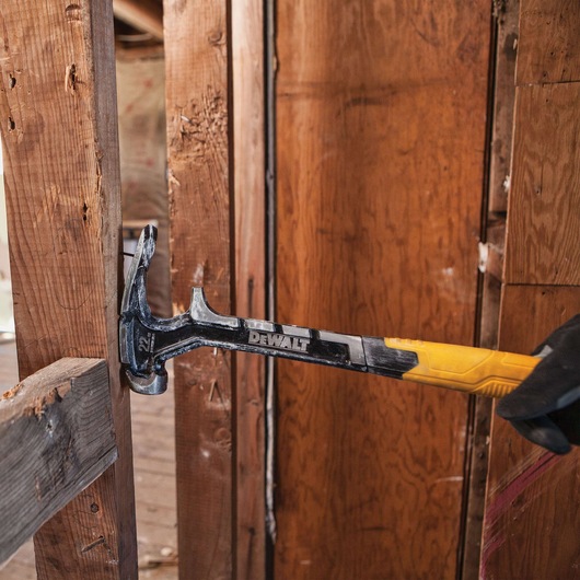 A DEWALT claw hammer with a yellow handle being used to remove a nail from a wooden beam inside a construction or renovation site.