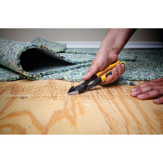 A close-up image of a hand using a yellow and black DEWALT cutting tool on a wooden surface, with carpet padding partially rolled back in the background.