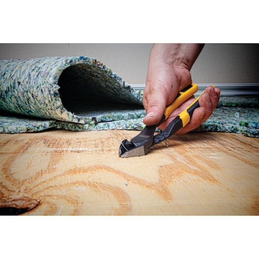 A close-up of a hand using DEWALT end cutting pliers (SKU: DWHT74274) to pull out a staple or nail from a wooden subfloor, with a rolled-up carpet pad in the background.