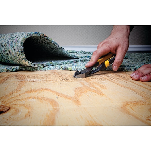 A person using a DEWALT hand tool to remove staples from a wooden floor with carpet padding partially lifted.