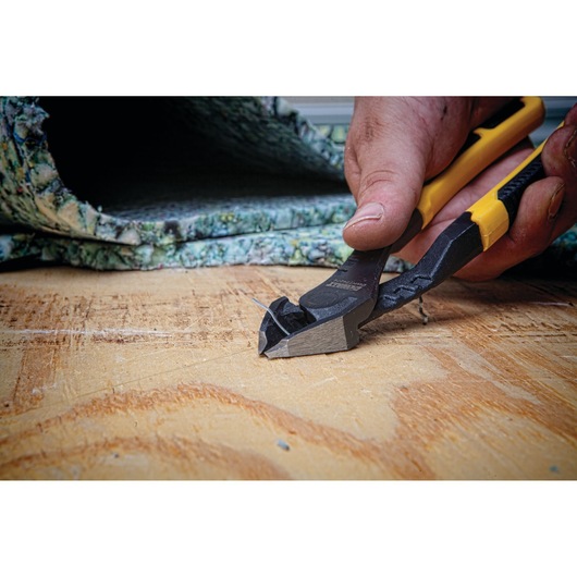 Close-up of a hand using a DEWALT wire cutter with yellow and black handles to cut a metal wire on a wooden surface. Carpet padding is visible in the background.