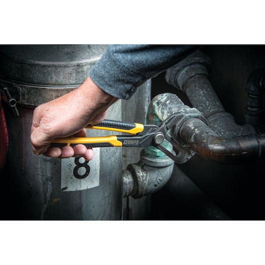 A person's hand using a DEWALT adjustable plier tool to grip a metal pipe in an industrial setting.