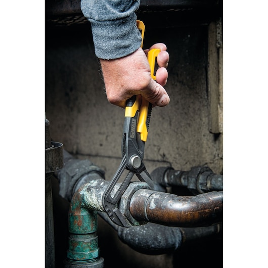 A person uses a DEWALT adjustable pliers tool with yellow and black handles to grip and turn a rusted metal pipe fitting in an industrial or plumbing setting.