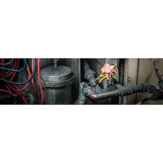A hand using a DEWALT adjustable wrench to tighten or loosen a pipe fitting in an industrial plumbing setting with several metal pipes and hoses visible.