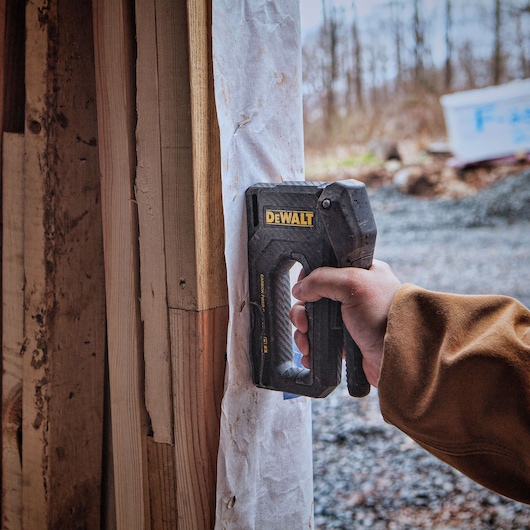 A person uses a DEWALT hand stapler to attach a sheet of material to a wooden frame at a construction site outdoors.