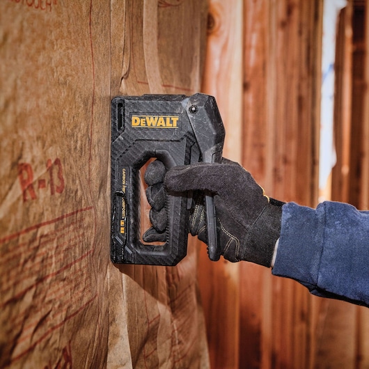 A person wearing black gloves using a DEWALT staple gun to secure insulation material to a wooden wall.