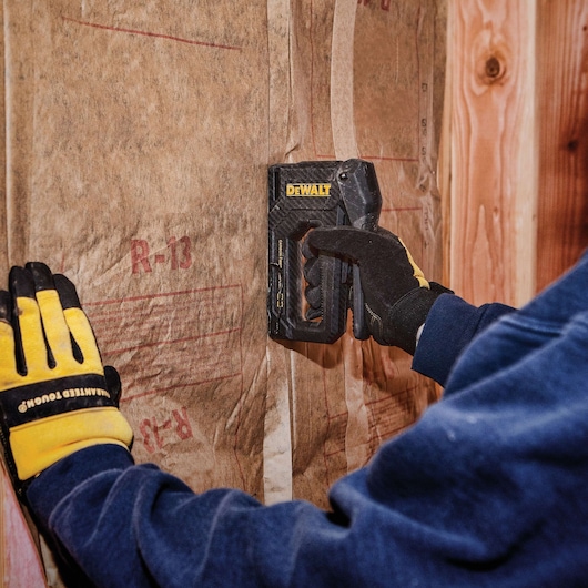 A person wearing work gloves uses a DEWALT stapler to secure insulation material to a wooden wall frame.