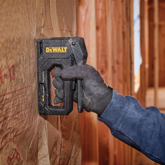 A person wearing a dark glove uses a DEWALT staple gun to attach insulation material to a wooden wall frame.