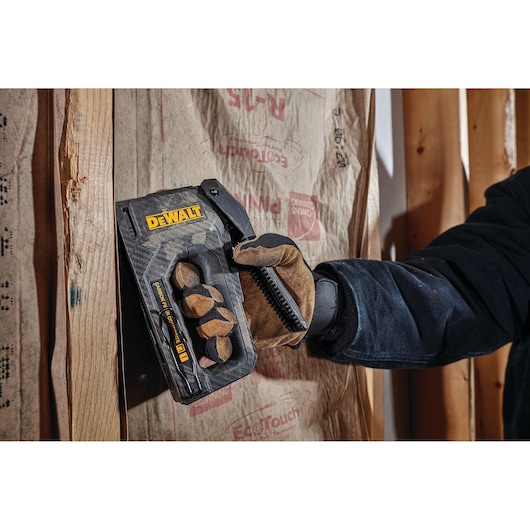 A person wearing work gloves uses a DEWALT stapler to attach insulation material to a wooden wall frame.