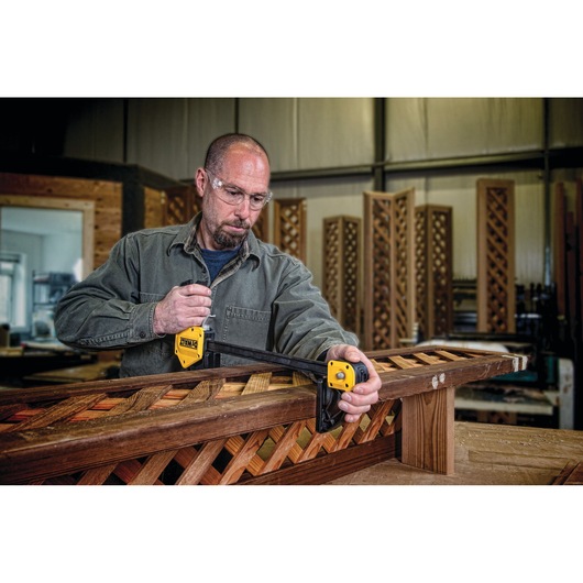 A person in a workshop uses a DEWALT DWHT83186 photography clamp to secure wooden lattice panels on a workbench. The person's face is intentionally blurred for privacy.