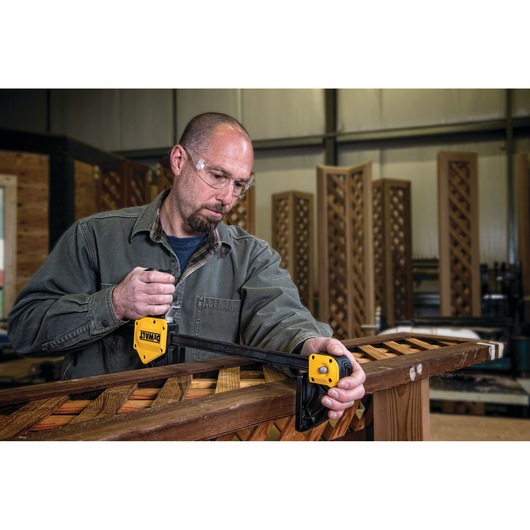 A person is using a DEWALT bar clamp (SKU: DWHT83186) to secure a piece of wooden lattice in a workshop. The individual's face is blurred for privacy.