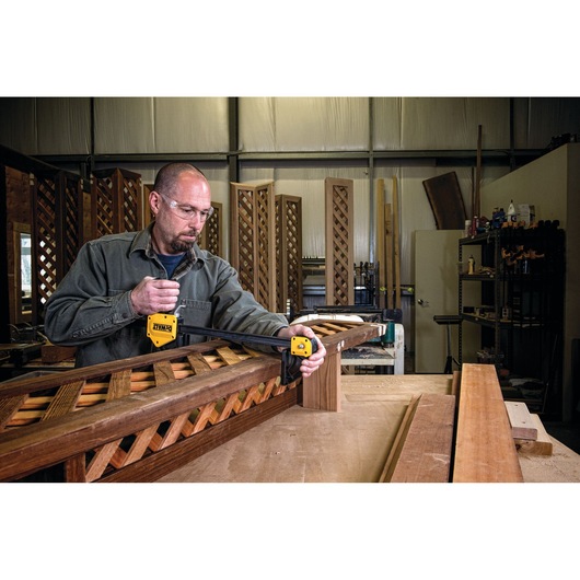 A person using a DEWALT bar clamp (model DWHT83186) to secure a wooden lattice panel on a workbench in a woodworking workshop. Various woodworking tools and materials are visible in the background.
