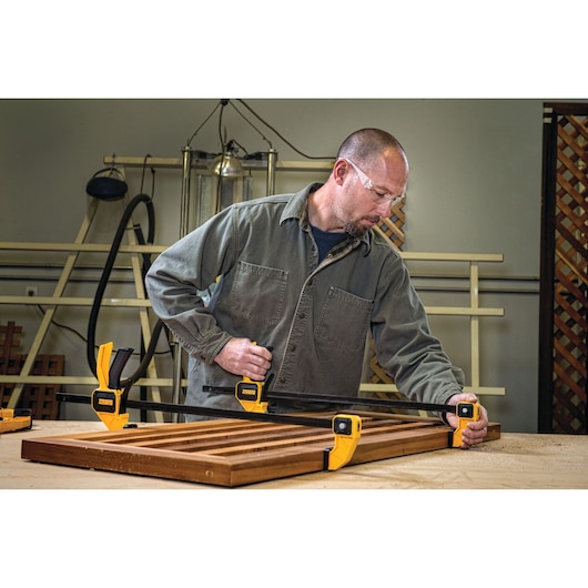 A person using yellow DEWALT clamps to secure a wooden frame on a workbench in a workshop. The person's face is blurred and the clamps are positioned at the corners of the wood.