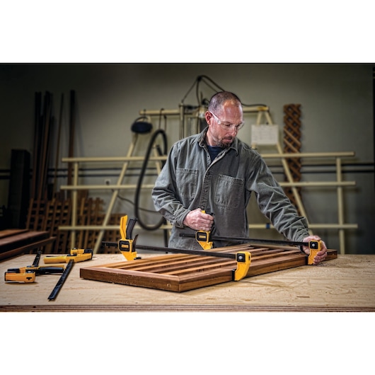 A person in a workshop uses yellow DEWALT clamps to secure a wooden frame on a workbench. Various woodworking tools and materials are visible in the background.