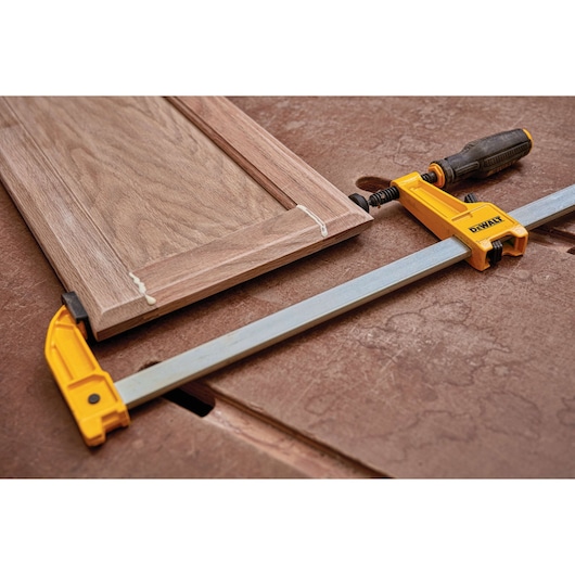 A DEWALT bar clamp (model DWHT83265) securing a wooden cabinet door on a workbench. The clamp has yellow and black handles and is used for woodworking projects.