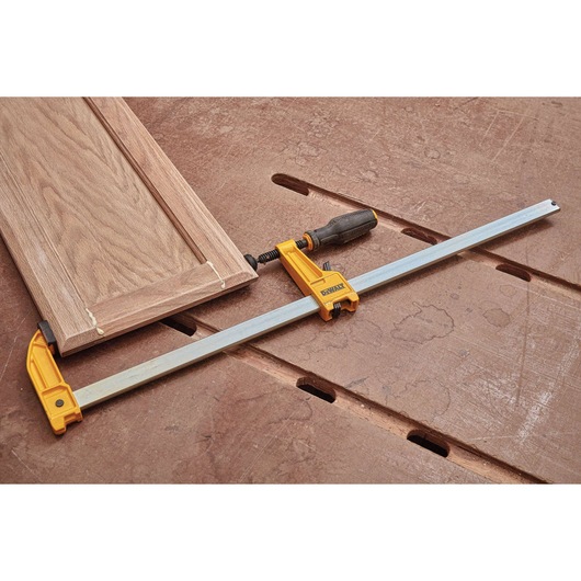 A DEWALT bar clamp is shown securing a wooden cabinet door on a workbench, highlighting its use in woodworking projects.