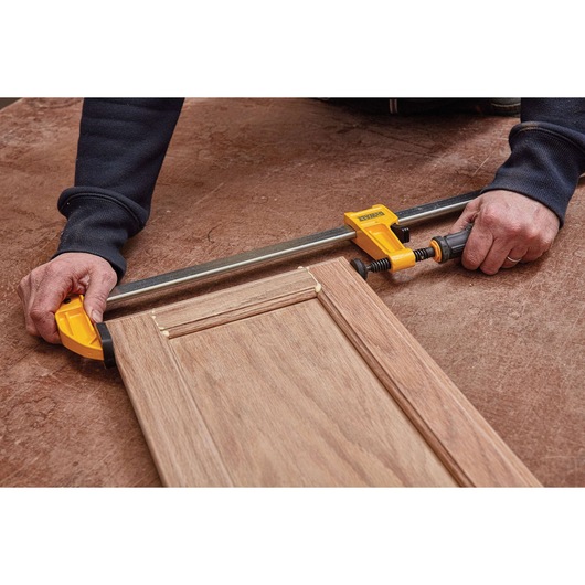A DEWALT yellow bar clamp is being used by a person to secure a wooden cabinet door on a work surface.