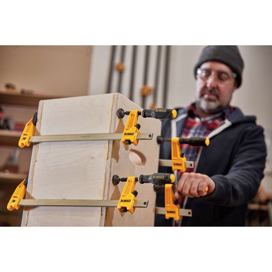 A person in a workshop uses four DEWALT bar clamps (model DWHT83829) to secure wooden boards, assembling a wooden box. The face of the person is blurred.