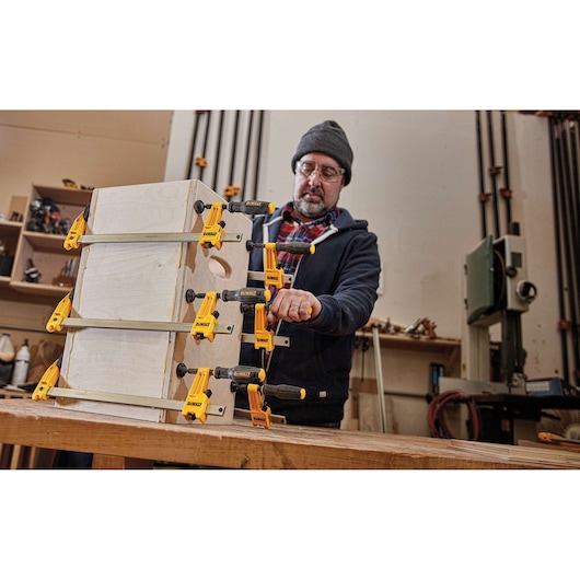 A person in a workshop uses several yellow DEWALT clamps (SKU: DWHT83830) to secure wooden boards together on a workbench.