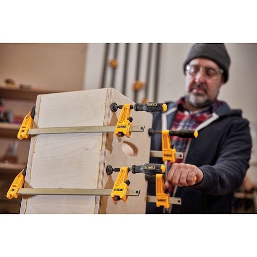 A person using DEWALT DWHT83830 bar clamps to secure two pieces of wood together in a workshop. The clamps are yellow and black, and the person's face is blurred.
