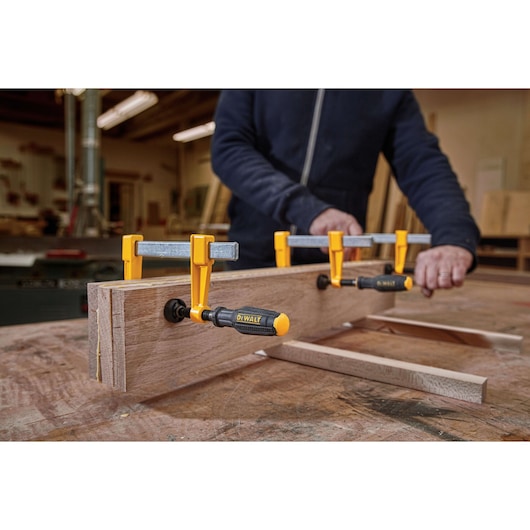 A person is working in a workshop, using several DEWALT clamps (SKU: DWHT83849) to secure pieces of wood on a workbench.