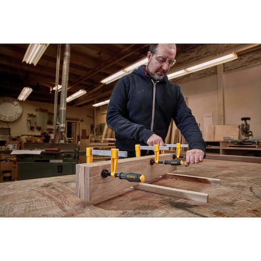 A person in a workshop uses DEWALT bar clamps (SKU: DWHT83849) to secure wooden boards on a workbench. The clamps have yellow and black accents and are holding the wood tightly for a woodworking project.