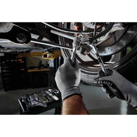 A person wearing gray gloves uses DEWALT hand tools to work on a car's suspension or brake system. A set of tools is visible on a table in the background.