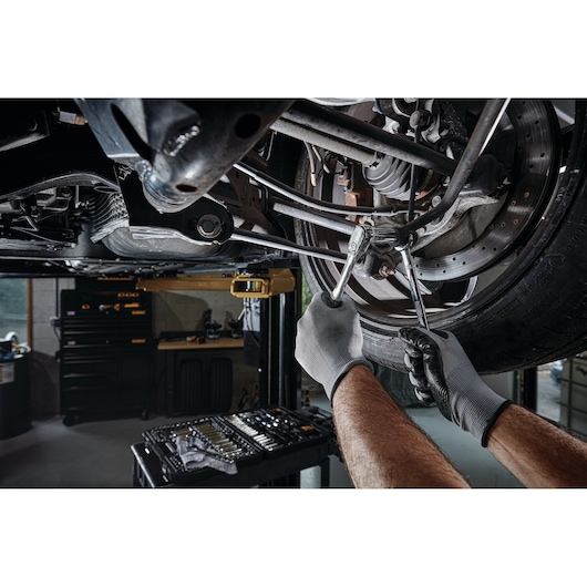 A person wearing work gloves uses wrenches to repair the suspension of a car, which is lifted on a hydraulic lift. A DEWALT mechanic tool set is visible in the foreground.