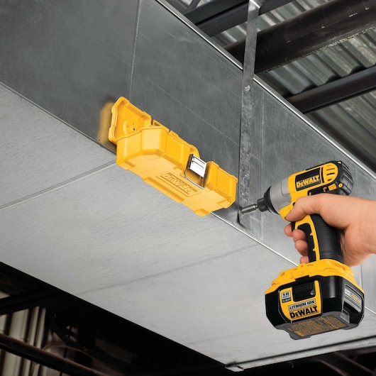 A person uses a yellow DEWALT cordless drill to attach a yellow DEWALT tool case to a metal duct or surface. The setting appears to be an industrial or construction environment.