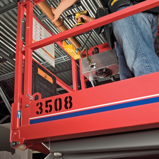 A person using a DEWALT cordless drill to work on a copper pipe while standing on a red elevated work platform marked with the number 3508. The scene is indoors with visible ceiling structures.