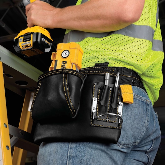 A close-up view of a person wearing a black leather tool belt with yellow stitching, containing various DEWALT tools and accessories. The person is also holding a DEWALT cordless power tool and is dressed in blue jeans and a bright yellow safety vest. A yellow ladder is visible in the background.