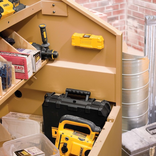 A close-up view of a tan-colored storage cabinet filled with various DEWALT tools and equipment, including a yellow drill, a black and yellow tool case, a yellow tool box mounted on the wall, and assorted boxes and containers. The scene is set in a workshop with a brick wall and metal barrels visible in the background.