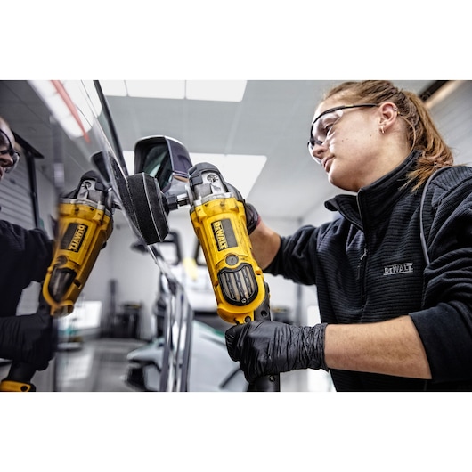 A person wearing black gloves and a black jacket is using a yellow DEWALT DWP849X polisher on the surface of a vehicle, with their face blurred.