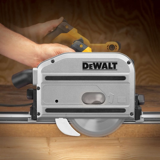 A close-up image of a DEWALT DWS520K track saw in use, cutting through a piece of wood on a guide rail. A person's hands are operating the saw, and the blade is visible spinning.