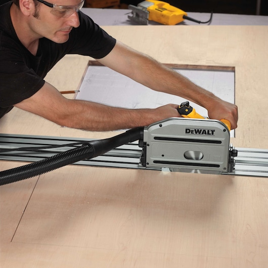 A person using a DEWALT DWS520K track saw to cut a sheet of wood, with the saw attached to a guide rail for precise cutting. The person is wearing safety glasses and the workspace includes additional tools and a vacuum hose connected to the saw.