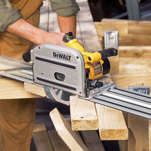 A DEWALT track saw being used to cut a wooden plank, with the saw mounted on a metal guide rail and operated by a person wearing a green shirt and brown pants.