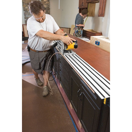 A person using a DEWALT DWS520K track saw to cut wood on a long work surface inside a kitchen under construction. Another person in the background is measuring cabinetry. The area features brown cabinets and a stone wall.