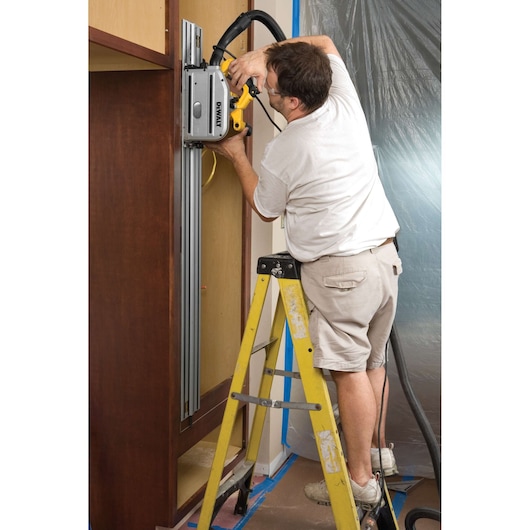 A person standing on a yellow ladder uses a DEWALT track saw (model DWS520K) to cut vertically along a wooden cabinet. The saw is attached to a guide rail and connected to a vacuum hose for dust extraction. The scene suggests indoor construction or cabinetry work.